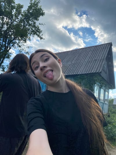 The image shows two people outdoors in what appears to be a rural setting, possibly Novyi Ural, Russia. A young person in the foreground is making a face at the camera, sticking their tongue out, with their head tilted sideways. They are wearing a black shirt. Behind them, another person, also wearing dark clothing, is visible from the shoulders up, with their head bowed.
In the background, an old, weathered wooden house with multiple windows is partially visible. The roof of the house is made of corrugated metal. Green foliage from trees and bushes surrounds the house and the people. The sky is cloudy with patches of blue, suggesting it might be daytime with mixed weather. The overall impression is casual and candid, likely a selfie or a photo taken by a friend. There is no visible text in the image.