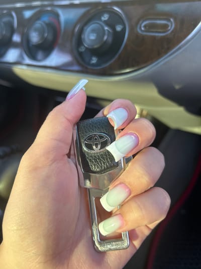 A close-up shot shows a person's hand holding a car key. The hand has long, almond-shaped nails painted with a French ombre design, transitioning from white at the tips to a pale pink at the cuticle. The car key appears to be for a Toyota, with the brand's logo visible on a black textured surface. The background is slightly blurred, but it seems to be inside a car, with part of the dashboard visible on the left. This includes what looks like a dial or button with wood-grain trim. The lighting suggests it's daytime, possibly outdoors, given the bright highlights. The focus is on the hand and the key, highlighting the polished nails. There is no visible text beyond the Toyota logo on the key.
