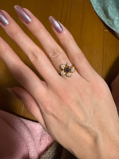 A close-up, overhead view depicts a light-skinned human left hand, oriented diagonally from the lower left to the upper right, showcasing a decorative ring on the fourth digit (ring finger). The hand's fingernails are long, with a pointed oval shape, and coated in a shimmering iridescent pink nail polish. The ring is gold-toned, featuring a multi-petal flower design composed of numerous small clear gemstones surrounding a central dark blue, possibly sapphire, cabochon or faceted gem. Visible in the lower left, a soft, light pink textured fabric is present, while the background consists of a polished brown wooden surface. The scene appears to be an indoor setting, specifically located in Kawasaki, Japan.