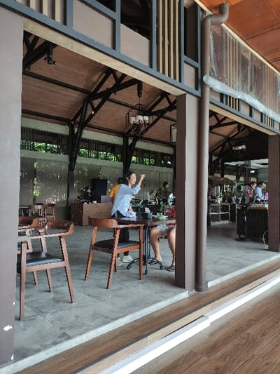 The image shows the interior of a restaurant located in Gambiran, Indonesia at the coordinates -8.434 latitude and 114.18 longitude. The scene is viewed from a low angle, looking up towards the ceiling. The ceiling is made of wood with a wooden frame. A woman with dark hair is standing on a chair, pointing upwards. She is wearing a light blue shirt and a yellow sweater. Next to her is a dark brown wooden table with two brown chairs. In the background are several other people sitting around tables, talking and eating. The restaurant floor is made of grey concrete. The restaurant has large windows, allowing for a view of the lush greenery outside. The overall tone of the image is relaxed and casual.