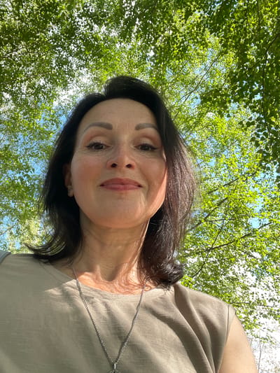 A woman with dark hair, wearing a beige t-shirt and a silver necklace, looks up at the camera from a low angle. She has makeup on, including eyeliner and lipstick. Her face is illuminated by sunlight, casting shadows on her skin. Behind her, green leaves and blue sky are visible, suggesting an outdoor setting, possibly under a tree. The image does not provide any specific details that would allow for identification of the city of Kirovo-Chepetsk, Russia.