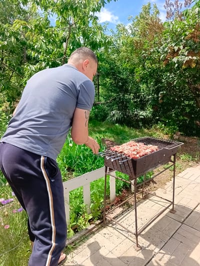 A man is grilling skewers of meat outdoors in a backyard. He is wearing a grey t-shirt and dark blue sweatpants with a white and brown stripe on the side. He has a tattoo on his left arm. The man is standing behind a dark metal grill, which is laden with raw meat on skewers. The grill is placed on a stand in a sunny outdoor setting, surrounded by lush green trees and vegetation. The ground is paved with concrete slabs, and there is a white railing to the left of the man. The sky is blue with scattered white clouds, suggesting it is daytime and the weather is pleasant. The scene appears to be in Vyazovka, Russia, indicating a residential or garden environment. The man's posture suggests he is actively involved in the grilling process, possibly preparing to turn the meat or tending to the fire. The overall mood is relaxed and domestic, typical of a summer day barbecue. There is no visible text in the image.