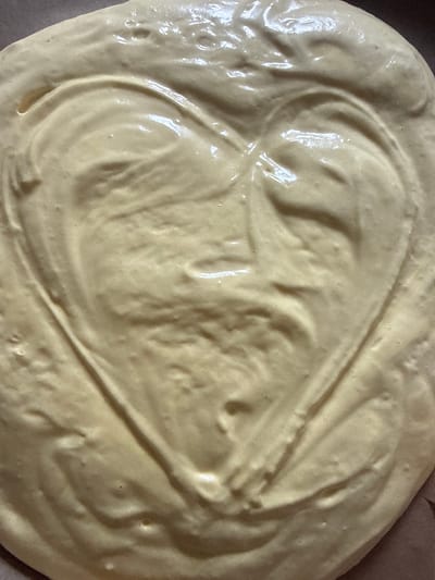 A close-up, overhead shot showcases a heart shape imprinted in a creamy, pale yellow batter, likely dough or cake batter. The batter has a smooth, glossy texture with visible ripples and indentations, suggesting it has been recently mixed or molded. The heart shape is formed by two curved lines meeting at a point at the bottom and forming two rounded lobes at the top. The lighting highlights the smooth, wet surface of the batter, creating subtle shadows and reflections that define the shape. The background appears to be a dark, flat surface, possibly a baking sheet or countertop. The image does not depict any specific location in the city of Hraniv, Ukraine, as it is a close-up of food preparation.