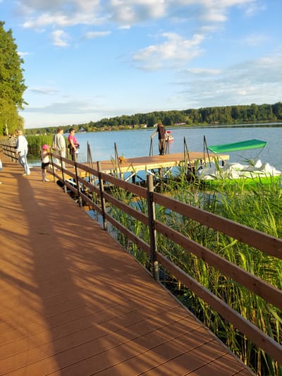 The image depicts a wooden boardwalk along a tranquil body of water, likely a lake or river, under a partly cloudy sky. The boardwalk, constructed from brown planks, runs diagonally from the lower left to the upper right of the frame. Several individuals are present on the boardwalk and a nearby pier. On the boardwalk, three women and a young child wearing a pink hat are walking or standing. A man in a dark shirt and pants stands on a wooden pier, leaning on a railing, facing away from the camera. Adjacent to the pier are two pedal boats, one white and green with a canopy, and another smaller red and white vessel further in the distance. Tall, green reeds grow along the water's edge, bordering the boardwalk and pier. The background features a dense line of green trees and houses, suggesting a semi-rural or suburban setting. The sunlight casts long shadows from the wooden railing onto the boardwalk, indicating it is late afternoon or early evening. This scene is located in the village of Razvetye, Russia.