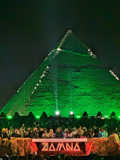 The image shows a night view of the Great Pyramid of Giza, illuminated with green light. A crowd of people is gathered to the right of the pyramid. Stage lights are visible, and a vertical sign reads "ZAMNA" in red neon. The sky is dark, and the surrounding area appears to be desert.