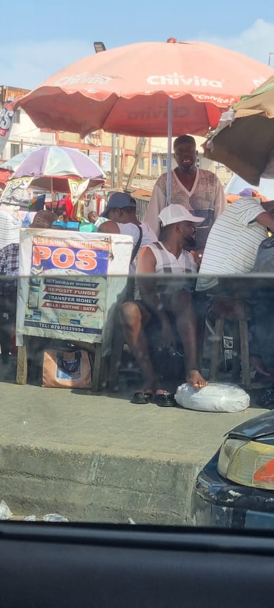A daytime street scene in Isunba, Nigeria, shows a roadside POS (Point of Sale) kiosk under a Chivita-branded umbrella. Several men are sitting and standing around the kiosk. One man has his bare foot resting on a wrapped object. A POS sign on the kiosk advertises services like money withdrawal, deposit, transfer, and bill/airtime/data purchase, with a visible phone number. A black vehicle is partially visible on the right.
