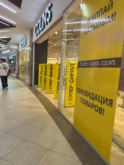 The image captures a street-level view of a shopping mall interior, specifically the storefront of a COLIN'S clothing store. The COLIN'S logo is prominently displayed in large, white, three-dimensional lettering on the store's facade. To the left of the COLIN'S store, a sign for "beautymania" is visible.
The COLIN'S storefront features large glass windows, offering a glimpse into the store's interior where clothing racks are present. Several large, yellow banners are displayed both inside and outside the store. One banner, partially obscured by the window, appears to have text in Cyrillic script, possibly indicating a sale or promotion. Another prominent yellow banner in the foreground features large, dark Cyrillic lettering that reads "ЛИКВИДАЦИЯ ТОВАРОВ!" which translates to "LIQUIDATION OF GOODS!". Above this, smaller text in white reads "COLIN'S COLIN'S COLIN'S". Another vertical yellow banner to the left also displays Cyrillic text and the COLIN'S logo at the bottom, repeating "СУПЕР ФИНАЛ" which translates to "SUPER FINAL".
A person is walking away from the camera down the tiled mall corridor, wearing a white t-shirt with graphics and black pants, and a black crossbody bag. The mall floor is tiled with light-colored square tiles. The lighting in the mall appears to be artificial, with overhead lights visible in the ceiling. Based on the context of the Cyrillic script and the presence of a COLIN'S store, this location is likely within Petropavl, Kazakhstan, as COLIN'S is a known brand with presence in Central Asia.