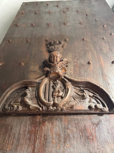 This is a close-up, eye-level, vertical shot of a highly detailed, carved wooden decoration on a large, dark wooden door. The door is constructed from planks with visible wood grain and is studded with numerous small, pointed metal rivets arranged in a grid pattern. The central decoration features a cherubic face at the top, wearing a crown. Below the face, an elaborate carving includes a shield or crest with vertical lines and a floral or leafy motif, surrounded by sculpted foliage and framing elements. The carving appears aged and weathered, with some areas showing signs of wear and damage. The overall impression is of an antique, possibly historical, architectural element. This decoration is located on the main entrance door of the Can Palau house in Mataró, Spain.