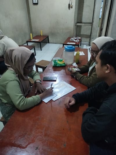 Three individuals are seated around a rustic, reddish-brown wooden table in an indoor eatery or casual restaurant, likely in Dramaga, Indonesia. The setting suggests evening or night due to the artificial lighting.
On the left side of the table, a woman wearing a light brown hijab, glasses, and a light green long-sleeved top is actively writing on papers with a pen. A black smartphone rests on the table near her. Opposite her, another woman wearing a white hijab and glasses is looking intently at a smartphone she holds in her hands. To her right, a man, seen in profile with dark hair and wearing a dark long-sleeved shirt, is observing the other two. Part of a fourth person in a light-colored hoodie is visible on the far left.
The table also holds a clear plastic water pitcher with a blue lid, a yellow squeeze bottle (possibly for sauce or condiments), and a green plastic bag, likely containing snacks. The background reveals simple, light-colored walls and a white tiled floor. Several other similar wooden tables with chairs are visible in the sparsely decorated room, each also featuring a yellow squeeze bottle. A clock is mounted on a distant wall, and some exposed electrical wiring is present. The mood appears focused, suggesting a casual meeting, discussion, or study session. No legible text is clearly visible on any objects.