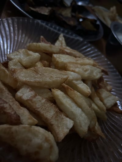 A close-up shot reveals a glass dish filled with thick-cut fries. The fries are golden brown, with a slightly uneven texture and some darker edges, suggesting they've been fried. The rippled glass surface of the bowl catches the light. Another bowl, partially visible on the left, contains a darker-colored food item, possibly roasted vegetables or burnt fries. The overall lighting in the image is dim, creating shadows and highlighting the texture of the fries and the glassware.