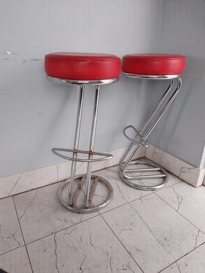 The image shows two red bar stools with chrome legs. The stools are positioned in a corner, with a light blue wall behind them and a tiled floor in front. The stools appear to be old and somewhat worn, with some rust visible on the chrome legs. The lighting suggests it is daytime, and the setting appears to be indoors, possibly in a home or a commercial establishment. There is no text visible in the image. The overall impression is one of a simple, utilitarian setting.