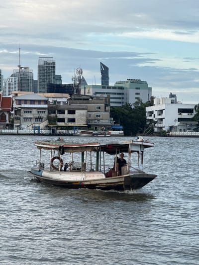 A medium shot shows a passenger boat with a single occupant navigating a wide, rippled river. The boat, white with blue and red accents, features a covered canopy supported by poles, and a life preserver is visible on its side. The occupant, a person wearing a dark shirt, stands at the helm. In the background, a dense cityscape unfolds along the riverbank. Buildings vary in style and height, ranging from older, low-rise structures with balconies and awnings to modern, tall skyscrapers with distinct architectural features. Lush green trees punctuate the urban landscape. The sky above is overcast with varying shades of gray and white clouds, indicating diffused daylight. The water is choppy, with visible wave patterns reflecting the sky. The scene appears to be set in Bang Kachao Subdistrict, Thailand, given the characteristic architecture and the presence of a river as a mode of transport in an urban setting.
