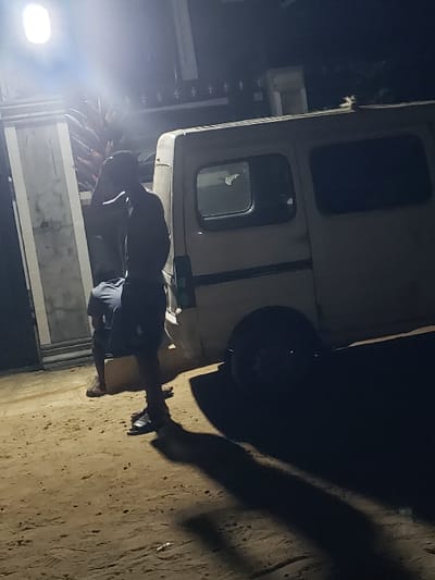 This nocturnal outdoor scene in Agunmo, Nigeria, depicts two individuals and a parked light-colored van on a dirt surface. A bright, rectangular artificial light source, positioned high on the left, illuminates the immediate area and casts long shadows to the right. A male figure stands in the foreground, facing away from the camera, wearing shorts and sandals, with his right arm raised towards his head. Behind and to his left, a second male figure, also in shorts and sandals, is bent over or crouching low, oriented towards the ground. A white or light-colored utility van, equipped with multiple side windows, is parked to the right of the individuals. To the far left, a concrete building wall or gate structure, adorned with decorative finials along its top edge, is partially visible. Reddish foliage is discernible behind the light source and adjacent to the building.