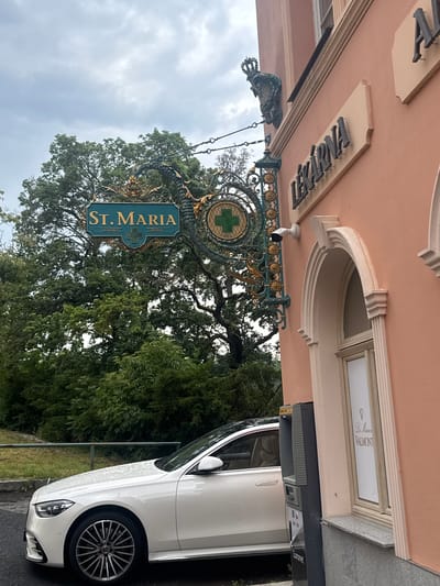 This is a medium shot, taken outdoors on an overcast day, of a building facade with a white car parked in front. The building is painted a peach color and features decorative architectural elements. Above an arched window, there is a sign that reads "Lékárna," indicating a pharmacy. To the left, an ornate hanging sign displays "ST. MARIA" in gold lettering on a teal background, with decorative dragon motifs and a green cross symbol. A white security camera is mounted on the wall near the hanging sign. The white car, a Mercedes-Benz S-Class, is parked on the street in front of the building. Trees and foliage are visible behind the hanging sign. An ATM machine is also visible to the right of the car. The overall scene suggests a street view in a town or city. Based on the "Lékárna" sign and the architectural style, this is likely Karlovy Vary, Czechia.