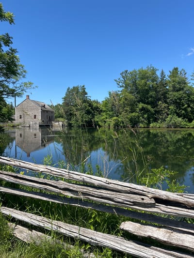 The image depicts a picturesque waterscape scene with a historic stone building reflected in the calm water. A weathered wooden fence runs along the right side of the frame, adding a rustic element to the composition. The building, likely a mill or similar heritage structure, features a stone facade and a multi-story design with visible windows and a chimney. Lush green trees and vegetation surround the water, creating a natural and serene backdrop. The reflection of the building and trees in the water is sharp and clear, suggesting a tranquil atmosphere. The sky above is a brilliant blue with a few wispy clouds, indicating fair weather and a sunny day. The overall scene evokes a sense of peace and historical charm, typical of rural settings. The photograph appears to be taken in the daytime, likely late morning or early afternoon given the bright sunlight. The location context provided places this scene in South Dundas, Canada, which is known for its natural beauty and historical sites. There is no visible text in the image.