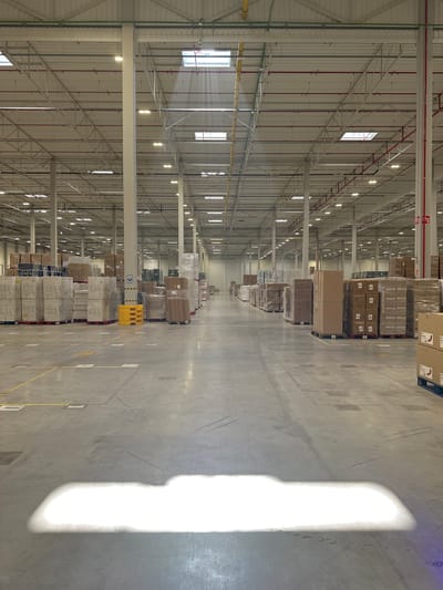 The image depicts the interior of a large, modern warehouse located in Sosnowiec, Poland. The high ceiling is supported by industrial steel beams and features numerous fluorescent lights and skylights, creating a well-lit environment. The floor is a smooth concrete surface, marked with yellow lines that appear to indicate pathways or storage zones.
The warehouse is filled with pallets of stacked boxes, many of which are wrapped in plastic shrink wrap, suggesting the storage of goods for distribution. Various types of boxes are visible, including cardboard boxes of different sizes and colors. In the mid-ground, a bright yellow industrial plastic pallet or bin is situated near a large white support column.
The overall impression is one of an organized and functional logistics facility. There are no people visible in the image, and the scene is static, focusing on the environment and the stored inventory. The lighting creates distinct shadows on the floor, adding depth to the scene.