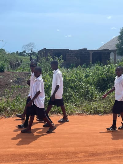 The image shows a group of four young boys walking along a dirt road in Ikoga, Nigeria. The boys are wearing white shirts and black shorts, which suggests they are likely students in school uniforms. They are walking in the same direction, casting shadows on the reddish-brown road. To the left of the road is a grassy area with dense vegetation and a building structure in the background with partially dilapidated brickwork. The sky is light blue with a few scattered clouds.