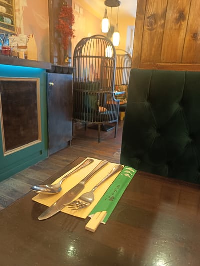 The image depicts a table setting in what appears to be a restaurant. On a dark wooden table, a place setting includes a spoon, a knife, and a fork, arranged on a yellow napkin. Next to the cutlery is a green paper wrapper with Japanese writing, likely for chopsticks, and a pair of wooden chopsticks. In the background, a plush, dark green upholstered chair is visible. Further back, the restaurant's interior features decorative birdcage-like structures, ambient lighting from pendant lamps, and wooden paneling. The scene suggests a contemporary dining environment, potentially with an Asian influence given the chopsticks wrapper. The location is identified as Killarney, Ireland.