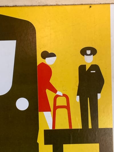 This is a close-up image of a graphic sign with a yellow background. On the left side of the sign, a large, dark brown, curved shape resembling the side of a vehicle is visible, with a white oval shape inside it. To the right of this, a stylized depiction of two figures is shown. The figure on the left is wearing a red top and white bottom, with a dark brown bun hairstyle. They are holding a red walker. The figure on the right is wearing a dark brown uniform with a white collar and hat, suggesting they are a uniformed officer or attendant. The figures are positioned on a dark brown platform with yellow accents. The image is a stylized graphic, not a photograph of a real-world scene. It is not possible to determine the specific location in Mineralnye Vody, Russia, from this image alone.