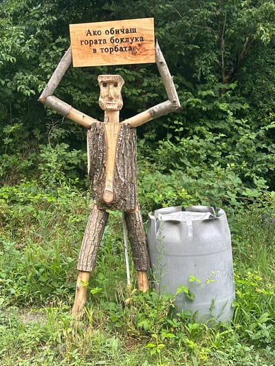 In Golyamo Belovo, Bulgaria, a whimsical wooden figure stands outdoors. The figure, crafted from logs and branches, holds a wooden sign above its head. The sign is inscribed with text in Cyrillic. To the right of the figure is a large, grey plastic barrel. The scene is set against a backdrop of dense green foliage, suggesting a natural or semi-rural environment. The lighting indicates it is daytime.