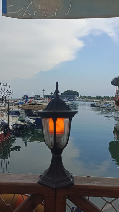 The image depicts a harbor scene in Mersin, Turkey, viewed from a seated perspective behind a wooden railing. In the foreground, a black outdoor lamp with a lit orange bulb is positioned centrally, its base resting on the railing. Beyond the lamp, the harbor is filled with numerous boats of various sizes, some appearing to be fishing vessels. The water is calm and reflects the sky and boats. In the background, a distant tree-covered island or peninsula is visible, along with more boats and structures along the shoreline. The sky is partly cloudy, with shades of blue and white. The overall impression is one of a quiet marina, likely during the early morning or late afternoon.