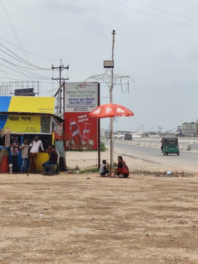 The image captures a roadside scene in Hatikumrul, Bangladesh. A makeshift shop with a yellow and blue facade is visible on the left, where several men are gathered. Adjacent to the shop is a large advertising sign for "National Food Village Highway Hotel & Restaurant," which includes the restaurant's name in both English and Bengali, along with contact information and drink advertisements. An orange "Polar" umbrella provides shade to a woman and a child who are sitting on the ground, potentially sharing a meal or snack. The immediate ground is dirt and gravel with scattered litter. In the background, there's a highway with visible construction work, vehicles, and a green auto-rickshaw moving on the road. Overhead, power lines and a light pole with tangled wires add to the busy landscape. The sky is overcast, suggesting a cloudy day.