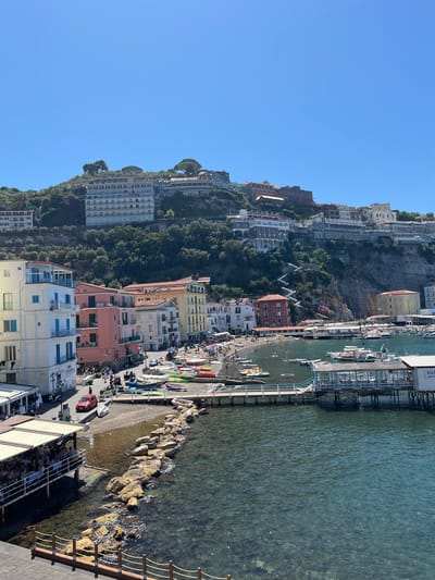This is a vertical, eye-level, medium shot of the waterfront in Sorrento, Italy, on a sunny day. The scene features a lively harbor area with a beach, buildings lining the shore, and boats in the water. The city is built on a cliff face, with many buildings cascading down towards the sea.
In the foreground, the clear turquoise water reveals the rocky seabed. A stone breakwater extends into the water, with a wooden pier on the right side of the frame. This pier has a covered structure with tables and chairs, labeled as "RISTO BAGNI," suggesting a restaurant and beach club.
Adjacent to the pier is a small, dark pebble beach populated with sunbathers and swimmers. Various small boats, including colorful kayaks and paddleboards, are pulled up on the beach or floating in the water. A few people are seen swimming or playing in the shallow areas of the sea.
Along the waterfront, a row of buildings in various pastel colors, including white, pink, and yellow, are tightly packed together. These buildings have multiple stories and balconies, with awnings extending over some of the ground-level shops. A red car is parked on the street in front of these buildings.
Further up the cliff, a multitude of buildings of varying sizes and architectural styles are visible, interspersed with dense green vegetation and trees. A prominent white, zig-zagging staircase is visible on the side of the cliff, leading up to the higher levels of the city. The sky above is a clear, cloudless blue.
The overall atmosphere is one of a busy, picturesque tourist destination, capturing the essence of a Mediterranean seaside town.