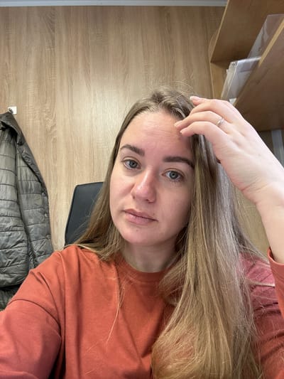 A woman with blonde hair and light complexion is visible in a selfie-style image. She is wearing a terracotta-colored long-sleeved shirt. Her right hand is raised, fingers touching her forehead. A silver ring is visible on her right ring finger.
The background includes a light brown wood-grain surface, a black chair, and a gray quilted jacket. Above the woman is a partially visible shelf.
There is no verifiable evidence of the specific location, Ural'skoe, Russia.