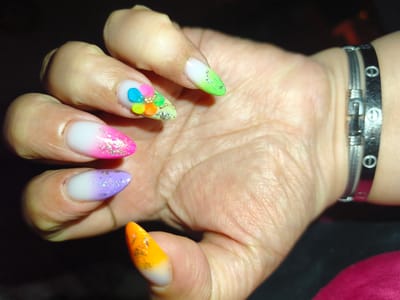 The image shows a close-up of a woman's hand with brightly colored, long, stiletto-shaped artificial nails. Each nail features a gradient of colors, such as pink and white, green and white, purple and white, and orange and white, often with glitter accents. The index finger nail has a small floral design with tiny colorful elements and gold embellishments. The hand is resting against a dark background, and a silver bracelet with a cable-like design and circular engravings is visible on the wrist. The lighting suggests an indoor setting, possibly a dimly lit room. The image does not contain any information that would place it in Komotini, Greece, or any other specific city.