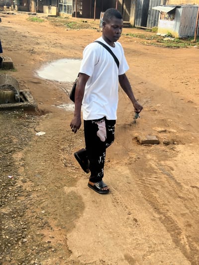 The image, oriented 90 degrees clockwise, captures a young man standing outdoors on an unpaved ground surface. The terrain consists of light brown packed dirt, sand, and scattered small gravel, marked by tire tracks and patches of lighter, possibly damp, soil or concrete.

The subject is a dark-skinned man with short hair, dressed in a white short-sleeved t-shirt and black trousers that feature white patterns, including visible text that appears to be "Summer." He wears black open-toed sandals, one of which displays a distinct logo resembling the Louis Vuitton emblem. A black strap, likely from a shoulder bag, crosses his chest. He holds a small, dark object, possibly keys or a device, in his right hand. His expression is neutral as he looks slightly downwards or to the side.

The background reveals a semi-urban or informal setting with various structures, including buildings constructed with corrugated metal sheets. Some sparse green vegetation is visible amidst the structures. In the lower left foreground, a concrete culvert or drain system is partially present.

The scene is brightly illuminated by natural sunlight, indicating clear, daytime weather, possibly late morning or afternoon, judging by the shadows. The man appears to be pausing or walking along the path. The visual cues, such as the building types and unpaved ground, are consistent with the provided location context of Ijegun, Nigeria.