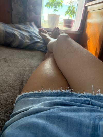 An indoor shot features a person's legs and feet as they lie on a bed in front of a window. The individual is wearing denim shorts with a frayed hem. Their feet, with painted toenails, are visible near the window. The background shows a window with plants in pots and sunlight streaming in. The environment appears to be a bedroom. There is no information to suggest this location is in Jalguba, Russia.