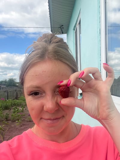 A young Caucasian woman with blonde hair tied in a messy bun is taking a selfie outdoors. She is wearing a bright pink sweatshirt. Her right eye is partially obscured by a ripe red strawberry, which she is holding with her right hand. Her fingernails are painted a vibrant pink. In the background, there is a light blue building with a white-framed window, a wooden fence, and a garden with green plants and some purple flowers. The sky is cloudy with patches of blue. The scene appears to be in a rural or suburban area, possibly in Сухринское, Russia, given the architectural style and vegetation.