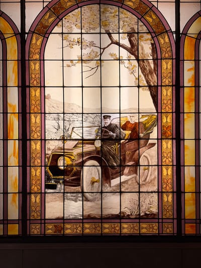 This is a vertically oriented image of a stained glass window, featuring a detailed pictorial scene framed by decorative glass panels.
The central scene depicts a man and a woman riding in a vintage open-top car. The man, presumably the chauffeur or driver, wears a cap and a dark coat and is positioned on the right side of the vehicle, holding the steering wheel. To his left, a woman with a bonnet and a warm-looking jacket sits as a passenger. The car itself is an early 20th-century model, characterized by its open design and classic details.
The setting appears to be outdoors, with a prominent tree branch extending into the left side of the frame, adorned with light-colored blossoms or foliage, suggesting a spring or early autumn scene. The background is light and somewhat abstract, implying a path or road through a natural landscape. The overall mood conveyed by the figures and setting is one of leisurely travel.
The pictorial scene is encased within an elaborate stained glass border. This border features a wide band adorned with repeating floral and leaf motifs in shades of gold, orange, and brown, set against a muted purple or mauve background. Above and below this main decorative band are simpler horizontal strips of plain and textured glass in light yellow and amber tones.
The style of the figures and vehicle within the stained glass is illustrative, resembling a painting integrated into the glass medium. There is a faint, unconfirmed signature "P. SCHATT" visible on a small glass panel in the lower right portion of the main scene.