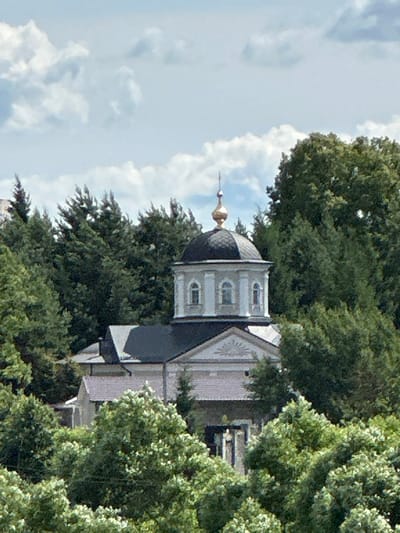 This is a vertical, medium-shot, slightly blurred photograph of the Church of the Sign of the Most Holy Theotokos in Dubrovitsy, Russia. The church's distinctive dome, with its dark grey cladding and golden spire, is visible peeking out from behind lush green trees. The building itself appears to be light-colored, possibly white or cream, with architectural details like arched windows and a decorative frieze visible beneath the dome. The surrounding trees are dense and full, their leaves a vibrant green, suggesting a warm season, likely summer or late spring. A bright, partly cloudy sky forms the backdrop, with fluffy white clouds scattered across a pale blue expanse. The overall lighting suggests it is daytime, perhaps late morning or early afternoon. The image focuses on the architectural elements of the church and its integration with the natural environment. There is no visible text in the image. The photograph does not appear to depict any people or specific activity, but rather a static view of the historical and religious site.