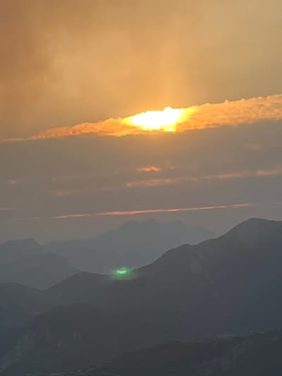 The image captures a mountain landscape at dusk or dawn, with the sun partially obscured by a horizontal band of clouds. The sky is a warm orange and yellow hue, transitioning to a muted gray towards the horizon. Several layers of mountains are visible, fading into the distance with increasing atmospheric haze. In the mid-ground, a prominent bright green light is visible on the slope of one of the mountains, appearing to emanate from a concentrated point. The overall scene is serene and natural, with the striking green light serving as a focal point against the subdued tones of the mountain range and sky. This phenomenon is known as a "Green Flash" and is a rare optical event that occurs just as the sun sets or rises. The image is a photograph of this natural event occurring in the vicinity of San Juan de Plan, Spain.
