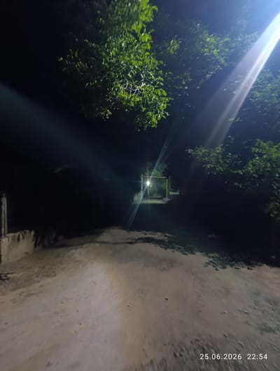The image depicts an outdoor scene at night, likely in a rural or semi-rural area, given the presence of a dirt or gravel road and vegetation. The time of day is indicated by the timestamp "25.06.2026 22:54" in the lower right corner, suggesting it is late evening.

The primary elements are a dirt road leading into the distance, flanked by dark trees and foliage. A bright light source, possibly a streetlamp or a strong flashlight, illuminates the scene from the upper right, casting long shadows and creating lens flare effects that streak across the image. In the background, a structure with a bright light in an opening is visible, suggesting it could be a gateway, entrance, or building.

The overall mood is somewhat mysterious due to the darkness and the bright, intrusive light. The presence of greenery suggests a temperate climate, and the timestamp points to summer. The image does not show any people or clear signs of specific activity beyond the illumination of a pathway. The location context of Slobozia-Dușca, Moldova, is provided, which may hint at the specific type of landscape or infrastructure visible.