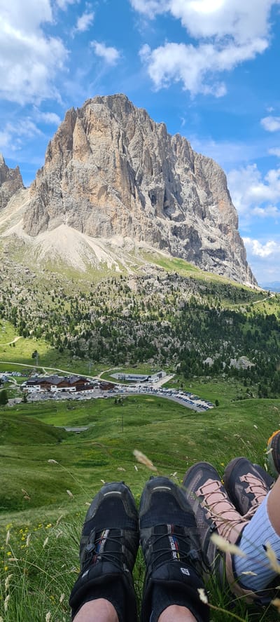The image shows a scenic view of the Dolomites in Val Gardena, Italy, with a prominent, rugged mountain peak dominating the background. The mountain is largely devoid of vegetation, displaying its rocky, eroded face under a bright blue sky with scattered white clouds. In the mid-ground, a green, rolling landscape is covered with sparse trees and bushes, leading down to a developed area with buildings, including a large lodge or hotel, and extensive parking lots filled with vehicles. A road curves through the area. The foreground features the lower legs and feet of two individuals sitting on a grassy slope, with their hiking boots visible. One pair of boots is black with red and yellow accents, and the other is a lighter color with pink laces. Blades of grass and some small yellow flowers are present in the immediate foreground. The overall scene suggests a recreational outdoor setting, likely a hiking or tourist destination.