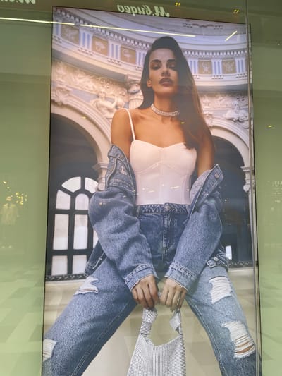 A large, brightly lit fashion advertisement featuring a woman is captured through a reflective surface, indicating it is likely displayed in a public space. The main subject is a woman with long, dark hair and striking makeup, posing with a confident gaze directly at the viewer.
She is dressed in a white strapless top with visible boning (resembling a corset or bustier), heavily distressed light blue denim jeans with prominent rips on the thighs, and an oversized blue denim jacket draped over her shoulders. Accessories include a multi-strand sparkly choker necklace and a small, rectangular sparkly silver handbag held by both hands at her waist.
The background of the advertisement features an ornate, classical architectural setting, characterized by light-colored stone columns, intricate carvings, and arched structures, suggesting a grand building or a studio backdrop designed to emulate one.
The reflective surface covering the advertisement shows various elements of the surrounding environment, including what appears to be a light green wall or paneling with subtle geometric patterns and numerous small, bright light sources reflecting from the ceiling or nearby fixtures.
Visible text within the advertisement, though partially obscured and rotated, includes "M.rindo" or "M. Kundo". Reflections also show inverted and fragmented text such as "YDE", "IIXPIII", "OJO", "video", and "0.0", suggesting other signage or displays in the vicinity. The image portrays a modern fashion campaign in what appears to be a commercial setting within Саларьево, Russia.