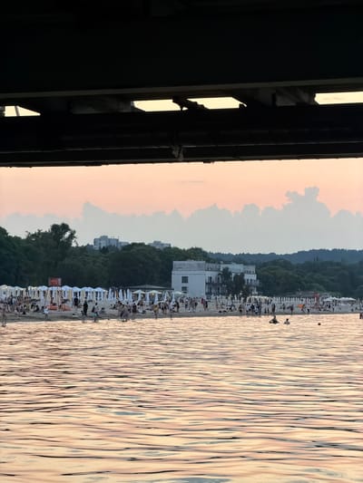 This is a photograph taken from under a bridge or pier, looking out towards a beach and the sea. The foreground is dominated by the textured surface of the water, reflecting the soft, pastel colors of the sunset sky. In the mid-ground, a crowded beach is visible, with numerous people engaged in various activities. Beach umbrellas and sun loungers are arranged in rows, indicating a popular recreational area. Several buildings, including what appears to be a hotel or apartment complex, are situated behind the beach, backed by a dense line of trees. The sky above is a soft gradient of pink and pale orange, with wispy clouds adding texture. The overall impression is one of a lively summer evening at a seaside resort. This scene is consistent with the waterfront of Sopot, Poland, a well-known spa town and seaside resort on the Baltic coast.