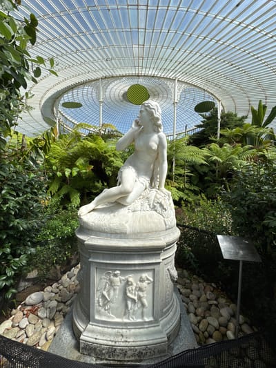 A white marble statue of a woman, identified by a nearby plaque as "Eve," is displayed on a pedestal within a greenhouse. The statue depicts Eve sitting on a rock formation, with one hand near her face. The pedestal is adorned with bas-relief carvings of human figures. The greenhouse has a glass roof and a white, ornate metal structure. Lush green ferns and other plants surround the statue. The scene is lit by natural sunlight filtering through the glass roof. A metal plaque with text is positioned to the right of the statue. The ground around the pedestal is covered in stones and mulch, with a black mesh barrier in the foreground. This greenhouse is located in the Glasgow Botanic Gardens, in the West End of Glasgow, Scotland, United Kingdom.