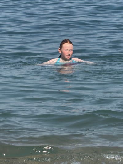 In a top-down perspective, a young person with fair skin and red hair is partially submerged in water. Their head is tilted back, eyes closed, and their mouth is open as if in mid-breath or exhalation. They are wearing a patterned swimsuit with blue and pink elements. The water is a calm, blue-grey, with distinct ripples and waves creating a textured surface. Sunlight glints off the water in the upper right portion of the image, suggesting a bright, possibly sunny day. The overall impression is one of peaceful immersion in the water. The provided crops offer closer views of the subject's face, hair, and swimsuit, as well as different sections of the rippling water.