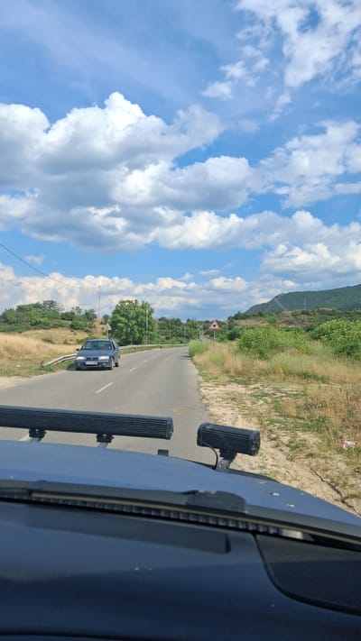 This is a full shot, eye-level, outdoor, daytime photograph taken from the perspective of a vehicle's dashboard. The vehicle is driving on a paved, two-lane road that curves to the right and heads toward a hilly, green landscape. The road is bordered by dry, grassy fields and scattered trees on both sides. In the distance, on the right side of the road, there is a triangular road sign with a black circle containing a smaller black circle. A blue four-door sedan is driving on the road in the opposite direction, approaching the viewer's vehicle. The sky is blue with scattered white, puffy clouds. The vehicle's dashboard is visible in the foreground, with two large, black auxiliary lights mounted on the roof. The scene is set in Mosomishte, Bulgaria.
