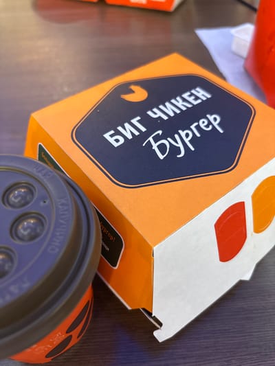 A close-up, angled shot captures a fast-food burger box and a coffee cup on a wooden surface. The burger box, predominantly orange with a dark blue hexagonal label, features white Cyrillic text that reads "Биг ЧИКЕН Бургер" (Big CHICKEN Burger). The coffee cup, dark gray with a brown lid, has partial Cyrillic text visible, including "КАПУЧИНО" (Cappuccino). The background is blurred, showing a hint of a red object and a white paper. The scene appears to be indoors, likely in a cafe or restaurant. No people are visible. The lighting is soft, suggesting an ambient indoor environment. The location of Shendzhii, Russia, is not visually ascertainable from the image.