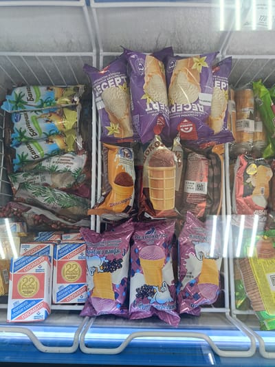 The image shows a freezer display of assorted ice cream products. The freezer is filled with various brands and flavors of ice cream, mostly in individual wrappers. Several packages feature prominent text in Cyrillic, indicating brands like "МОРОЖЕНОЕ", "ПЛОМБИР", and "ПЕТРОХОЛОД". Some packages depict images of ice cream cones, fruit, or cartoon characters. The products are arranged on shelves within the freezer, with some items stacked behind others. The lighting suggests the image was taken inside a commercial establishment. Without additional context, the specific location in Berdiansk, Ukraine, cannot be determined.