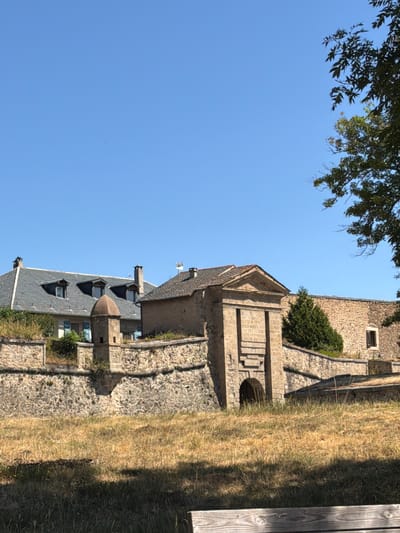 The image shows the ramparts and fortifications of the citadel of Mont-Louis, France, on a clear, sunny day. The fortifications are made of stone and feature an arched gateway. Behind the ramparts, a building with a grey tiled roof and dormer windows is visible, along with other stone structures. The foreground is dominated by dry, yellow grass. A tree with green leaves partially obscures the upper right corner of the image. The sky is a bright, cloudless blue. There are no people visible in the image.