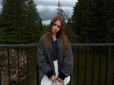 A young woman with long, brown hair stands on a bridge, looking at the camera with a neutral expression. She is wearing a dark blue crop top, a white pleated skirt, and a dark grey bomber jacket. She is holding a blue bag with both hands. Behind her, a body of water with reeds and debris is visible, separated by a black metal railing. The background is filled with dark green trees under a cloudy sky. The overall mood is somber, possibly due to the overcast weather. No text is visible in the image. The location is Chusovoy, Russia.