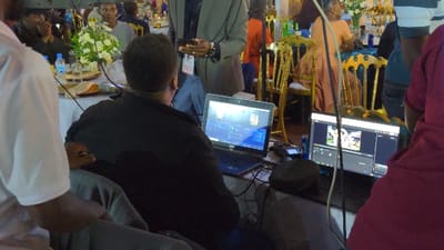 Two laptops are the focal point, both displaying video editing software. One shows a timeline, the other a preview. A person in a black shirt sits facing them, seemingly working on the project. A man in a suit stands nearby. The scene is cluttered, with tables, chairs, and people in the background. The event appears to be social, possibly a wedding reception, based on the floral arrangements and table settings. The overall impression is a behind-the-scenes glimpse of a video being edited at an event.