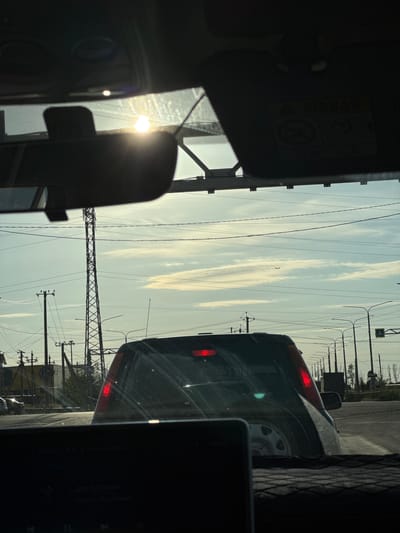 This media file is a photograph taken from inside a vehicle, looking out at a road scene. The perspective suggests the photographer is in the passenger seat, with the windshield and dashboard in the foreground. The sun is visible, partially obscured by the vehicle's interior, indicating it is either sunrise or sunset. The sky is a pale blue with wispy white clouds, suggesting fair weather.
The primary subjects are the road and other vehicles. A dark-colored SUV is in front of the photographer's vehicle, with its brake lights illuminated, suggesting it is either stopped or slowing down. There are numerous utility poles and power lines crisscrossing the sky, common in many urban and suburban areas. The landscape appears somewhat arid with sparse vegetation visible in the distance.
The setting is a road, likely a highway or a major road, given the presence of multiple lanes and the overhead power infrastructure. The presence of power lines and utility poles are strong location cues. The image depicts a common travel or commuting scene. There are no people clearly visible.
Notable details include the glare of the sun on the windshield, the red brake lights of the SUV, and the intricate network of power lines. There is some text visible on the side of the SUV in the distance that appears to be a logo or advertisement, but it is too blurry to read. A yellow sign with a black symbol is also visible on a pole. The overall mood is neutral, capturing a mundane moment of travel.
The file appears to be oriented vertically, suggesting it might have been captured by holding a phone in portrait mode or by rotating the camera. The location context of Novomar'evskaya, Russia, implies a setting in that geographical area.