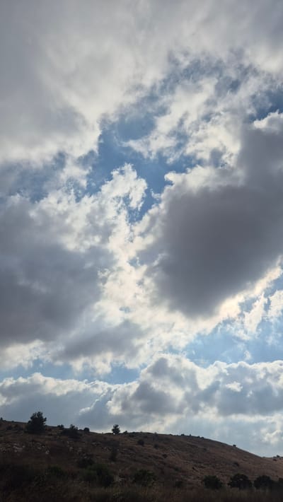 The image captures a vast sky filled with a mix of fluffy white and dark, heavy clouds. Patches of bright blue sky are visible between the clouds, suggesting a dynamic weather pattern. Below the dramatic sky, a rolling hillside is depicted. The terrain is covered in dry, golden-brown grass and sparse bushes, indicating a dry season. Scattered on the hillside are a few small trees with dark green foliage. The overall impression is a natural landscape under a partly cloudy sky. The specific location is Modiin-Maccabim-Reut, Israel, a city situated in a region known for its hilly terrain and Mediterranean climate.