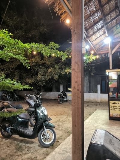 The image shows a nighttime scene in Cempeudak, Indonesia. Several motorcycles are parked on a sandy area beneath a tree with string lights. In the background, a building with a window is visible. A wooden post is in the foreground, and a food stall with the words "Mie Udang," "Mie Daging," and "Mie Telur" can be seen on the right side of the image. The area appears to be an outdoor dining space with a thatched roof.